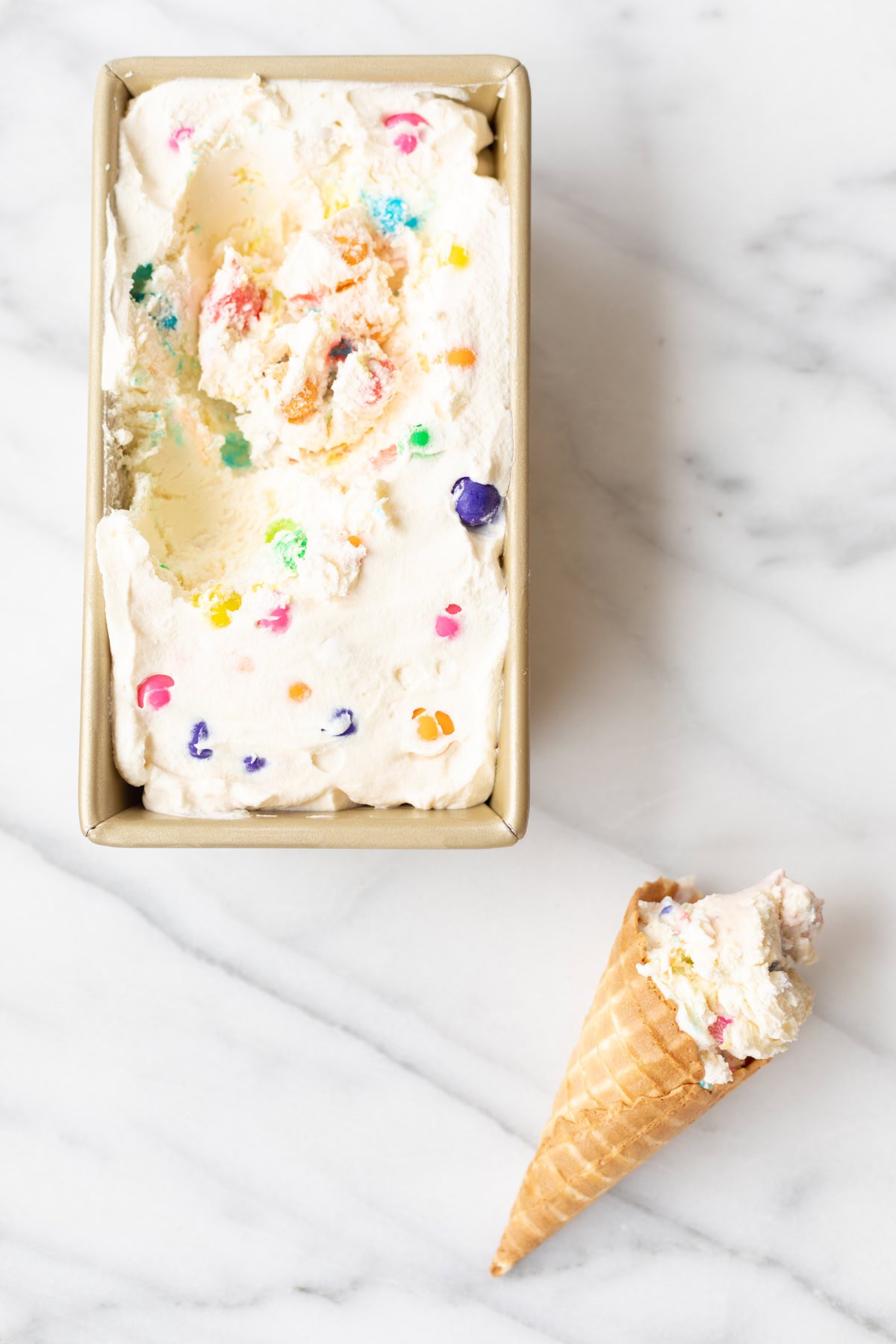 A rectangular container of colorful bubble gum ice cream sits on a marble surface with a scoop of the same ice cream in a waffle cone next to it.