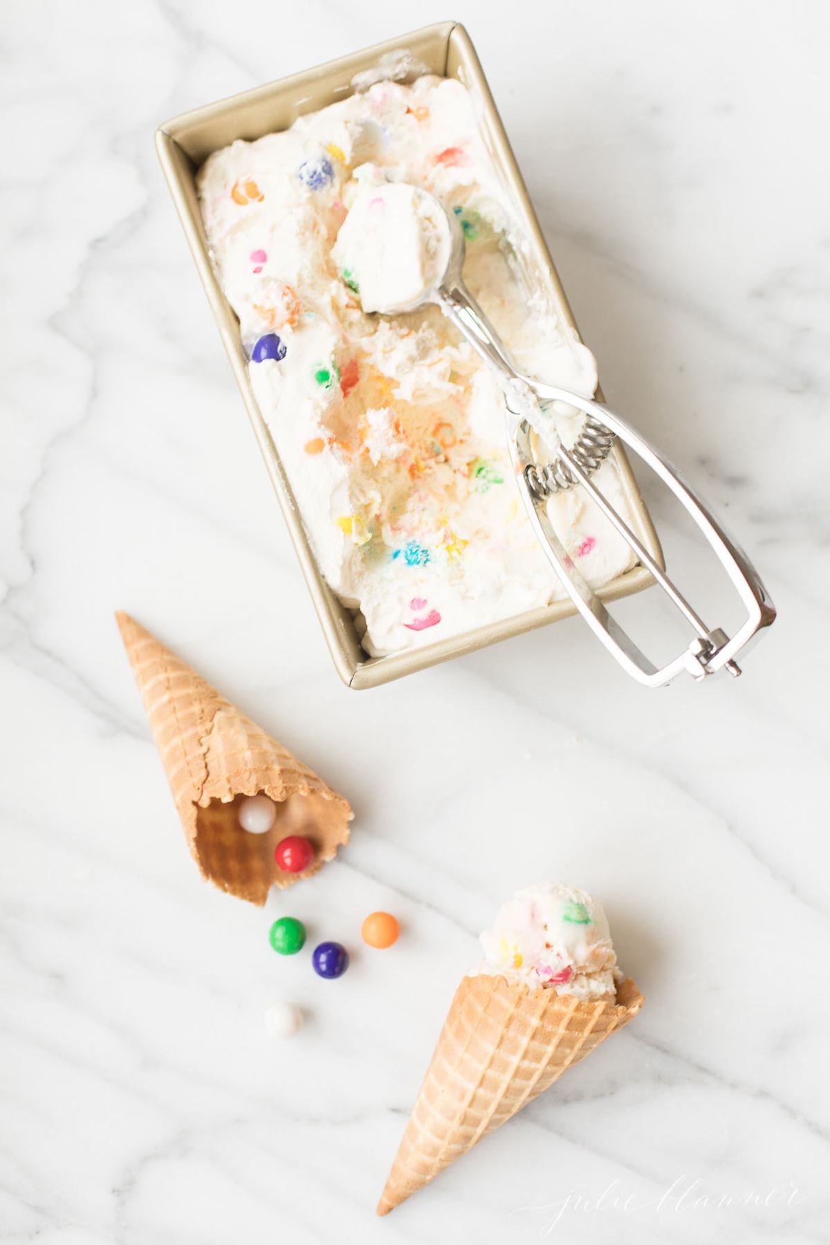 Bubble gum ice cream in a gold loaf pan, with cones nearby on a marble countertop. 