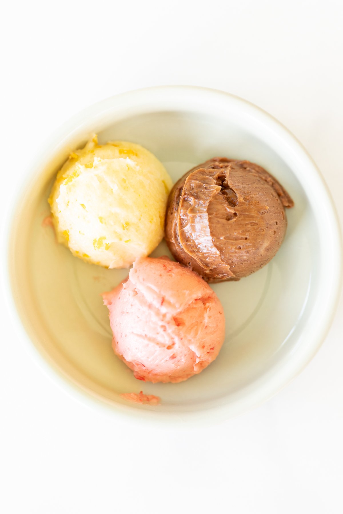 A small white bowl with a scoop of chocolate butter, strawberry butter and orange butter.