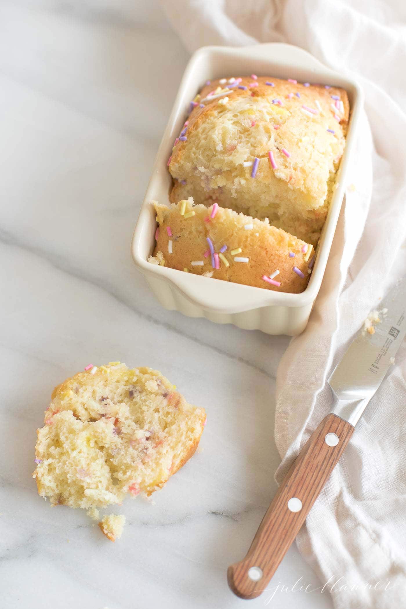 A funfetti bread recipe in a mini white loaf pan on a white marble surface.