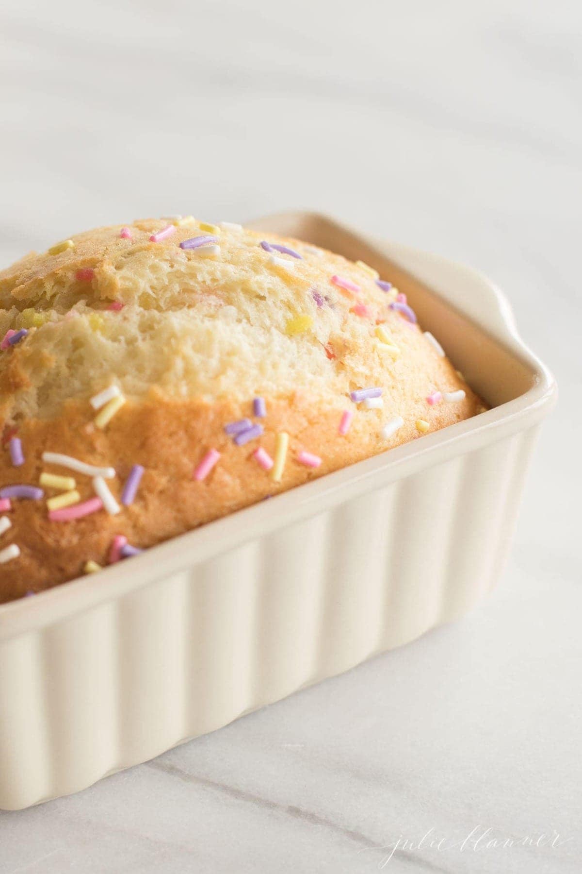 A funfetti bread recipe in a mini white loaf pan on a white marble surface.