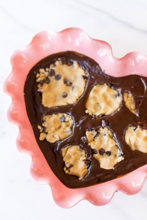 Heart-shaped baking dish filled with brookie batter mixed with dollops of cookie dough and sprinkled with chocolate chips.