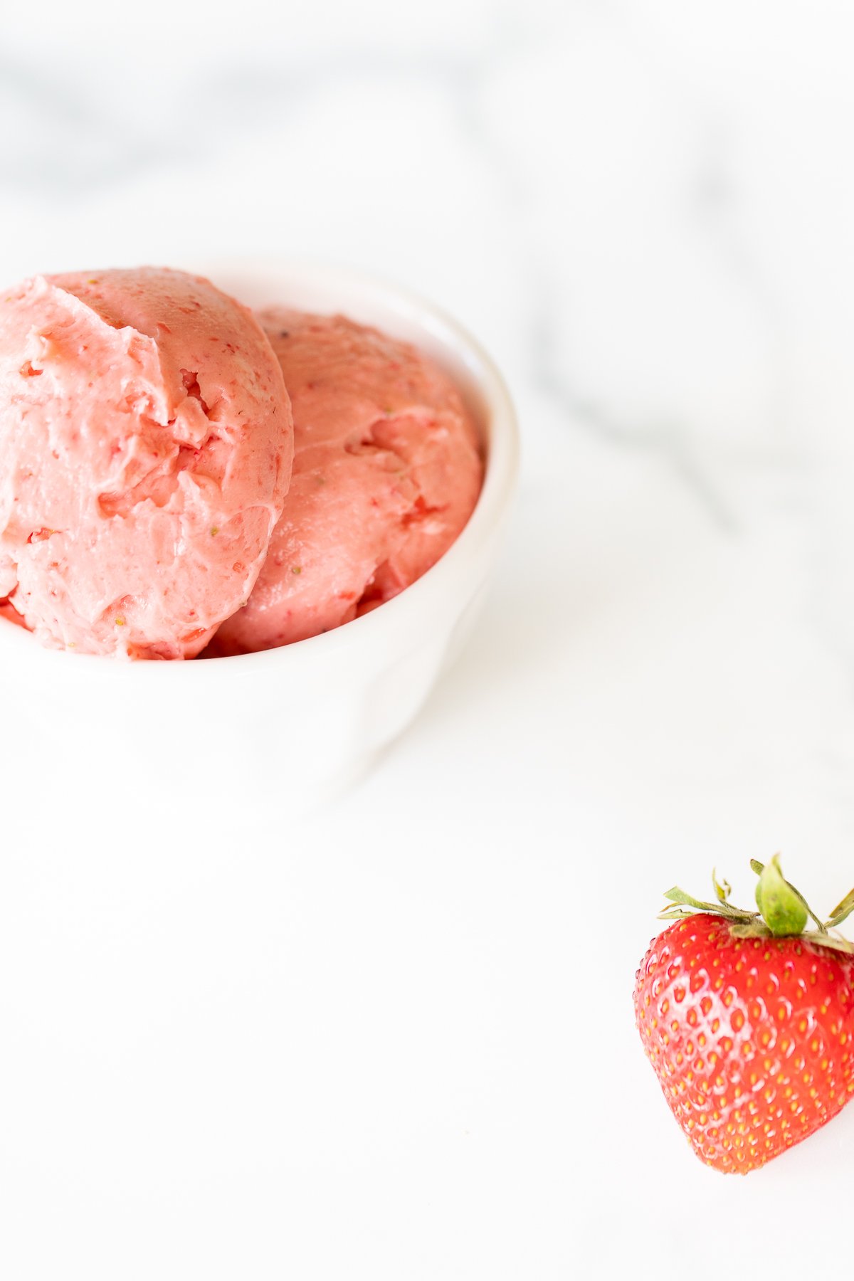 A small white bowl full of pink toned strawberry butter, on a marble surface.