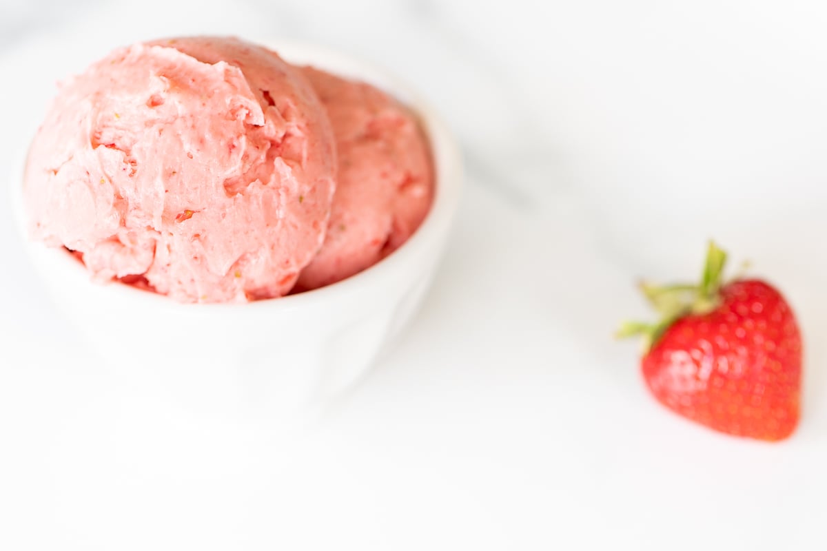 A small white bowl full of pink toned strawberry butter, on a marble surface.