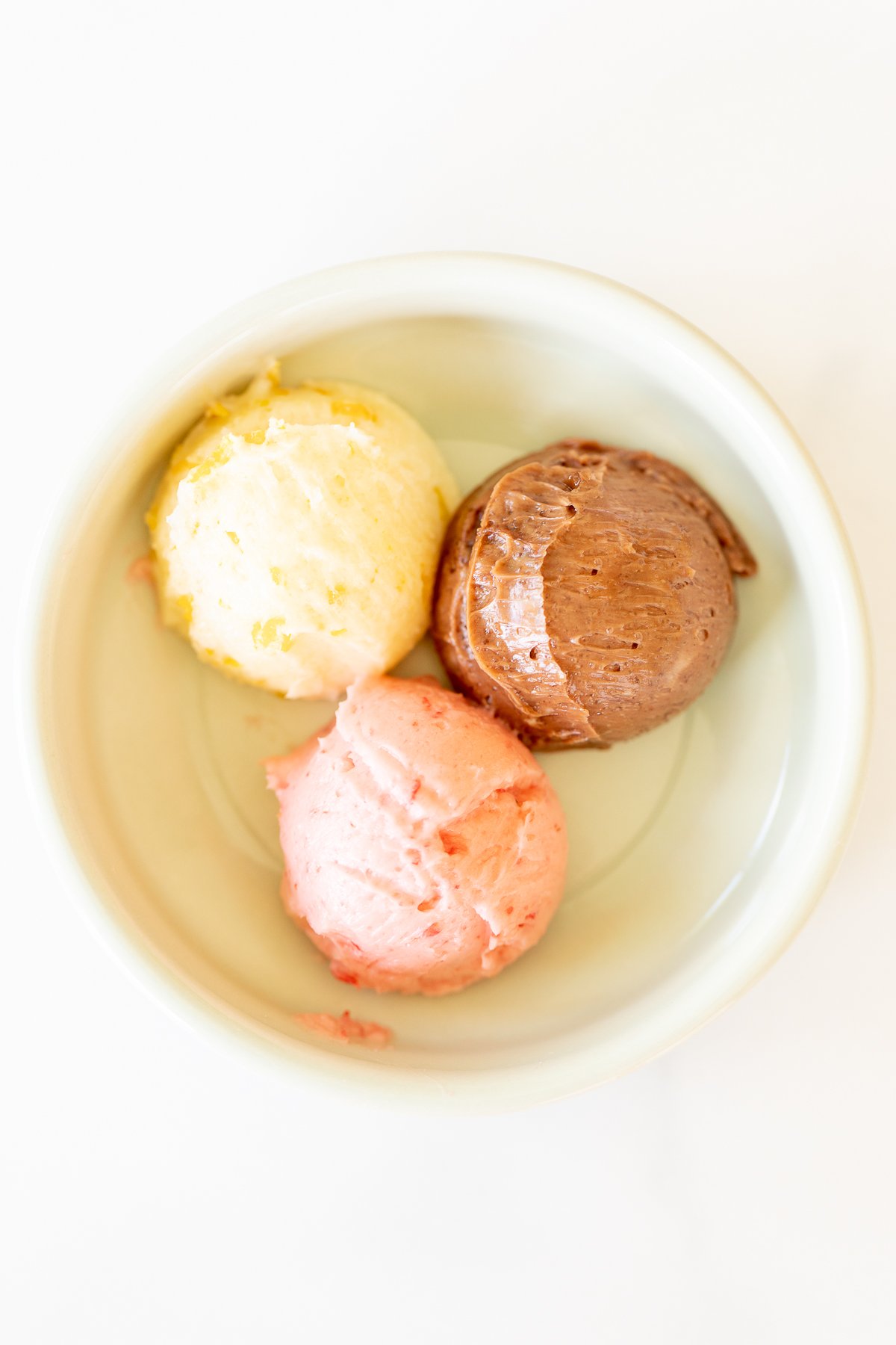 A small white bowl with a scoop of chocolate butter, strawberry butter and orange butter.