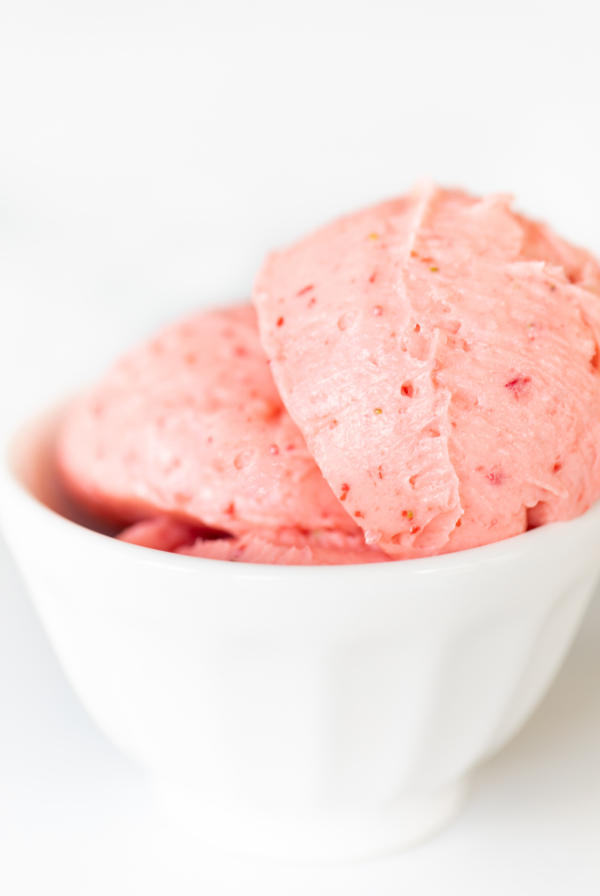 A small white bowl full of pink toned strawberry butter, on a marble surface.