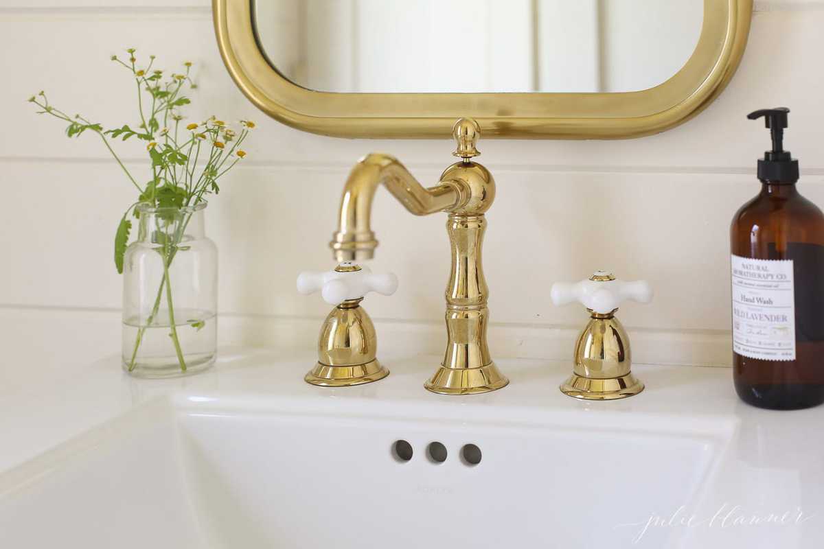 A white bathroom sink close-up shot, brass faucet and brass mirror above.