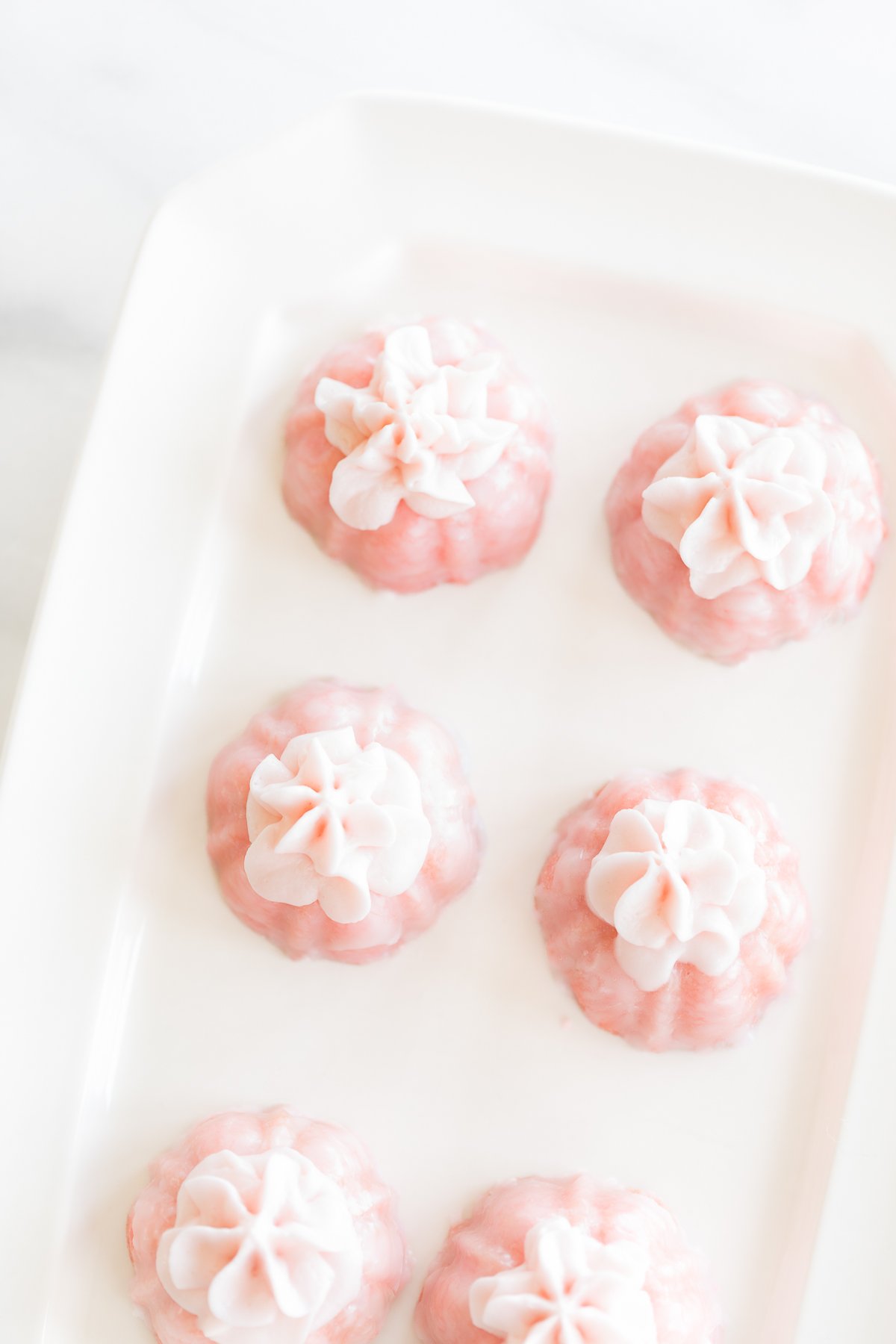 White tray with five pink champagne cupcakes, each topped with a white flower, arranged neatly on a marble surface.