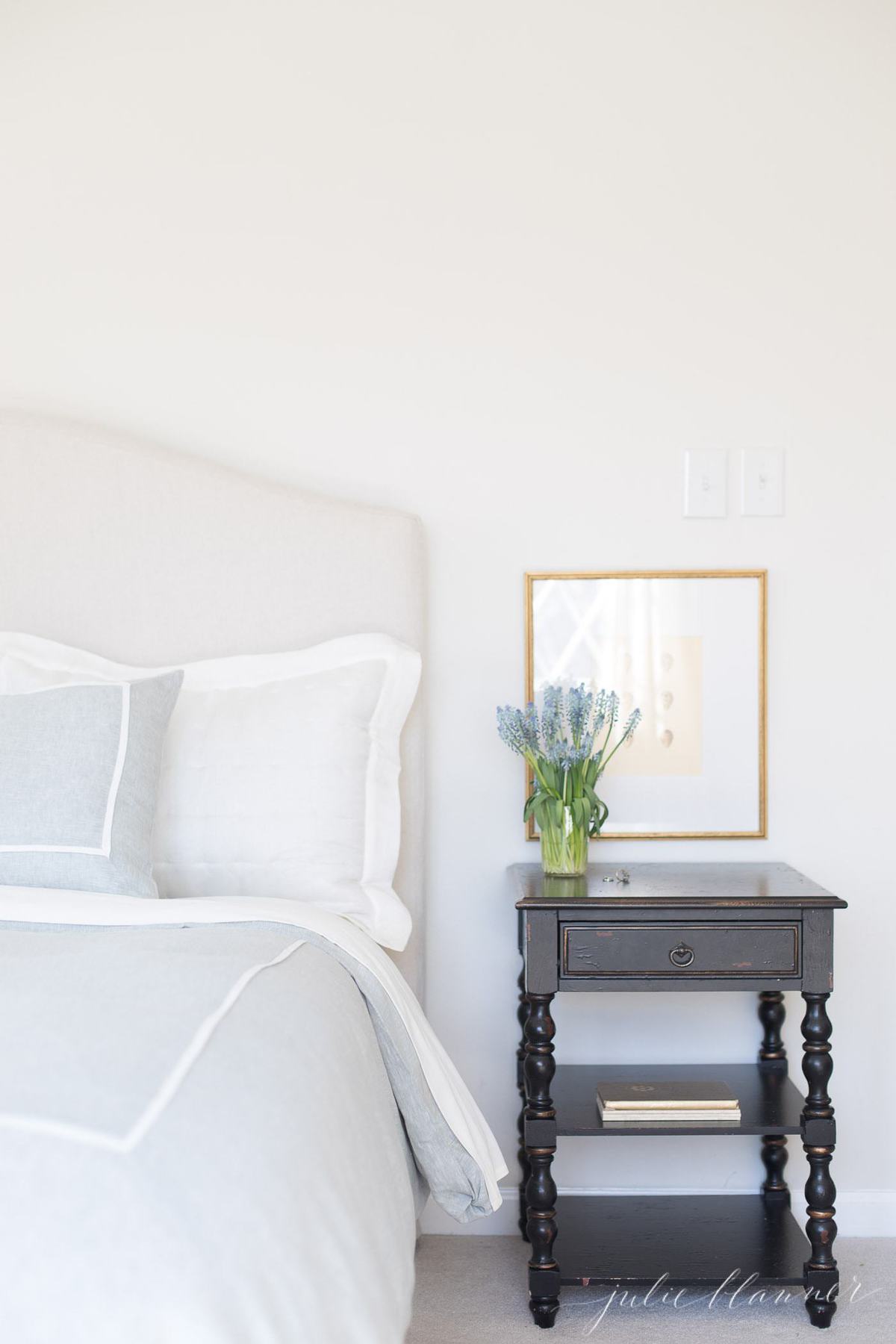 A white bedroom with an upholstered headboard and black nightstand, antique egg printable art over the nightstand.