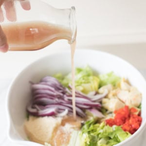 A hand is pouring red wine vinaigrette from a glass bottle into a large white bowl containing chopped vegetables, including lettuce, red onions, and red bell peppers.