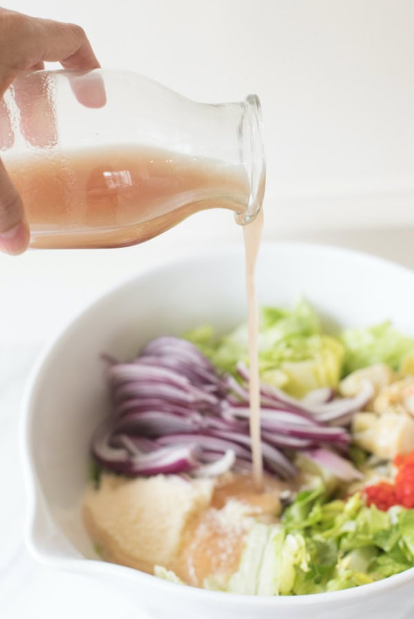 A hand pours a light brown red wine vinaigrette from a glass bottle into a bowl containing chopped lettuce, sliced red onions, and other salad ingredients.