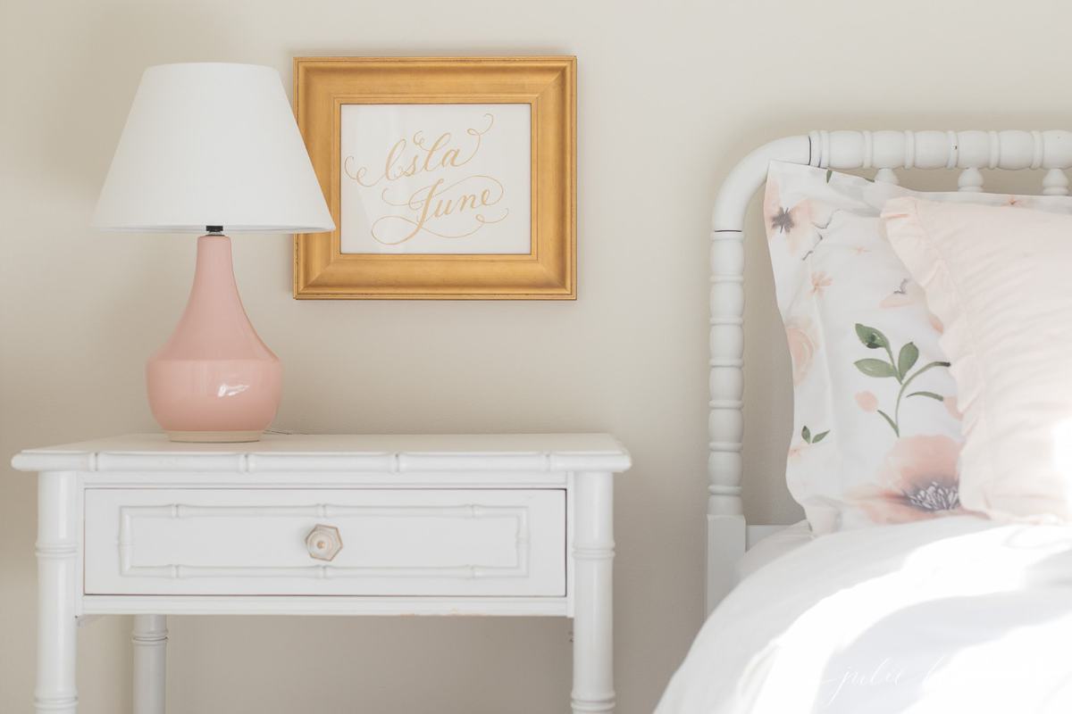 A white girl's bedroom with a pink lamp on a bedside table.