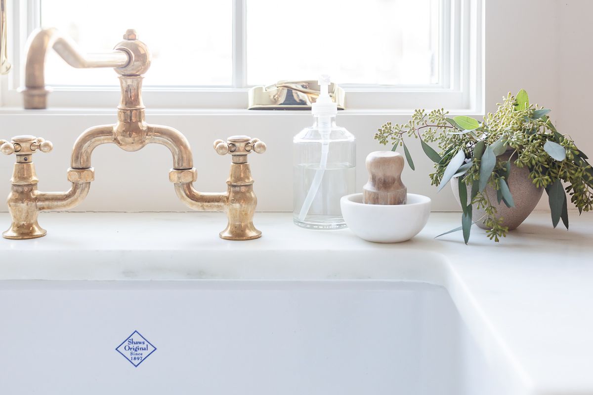 A white farm sink with a brass faucet and eucalytpus in a vase