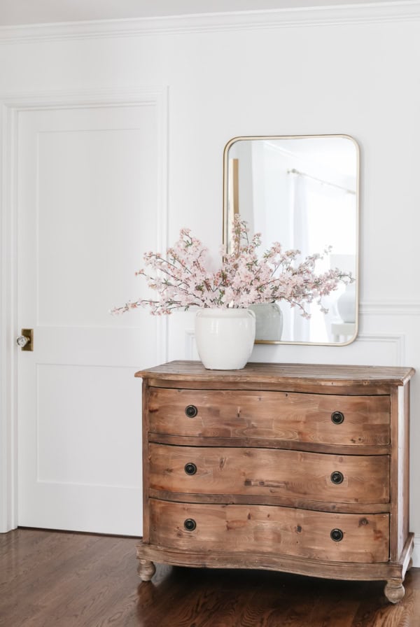A wooden dresser with three drawers stands against a white wall. A large mirror and a white vase with cherry blossom branches are placed on top.