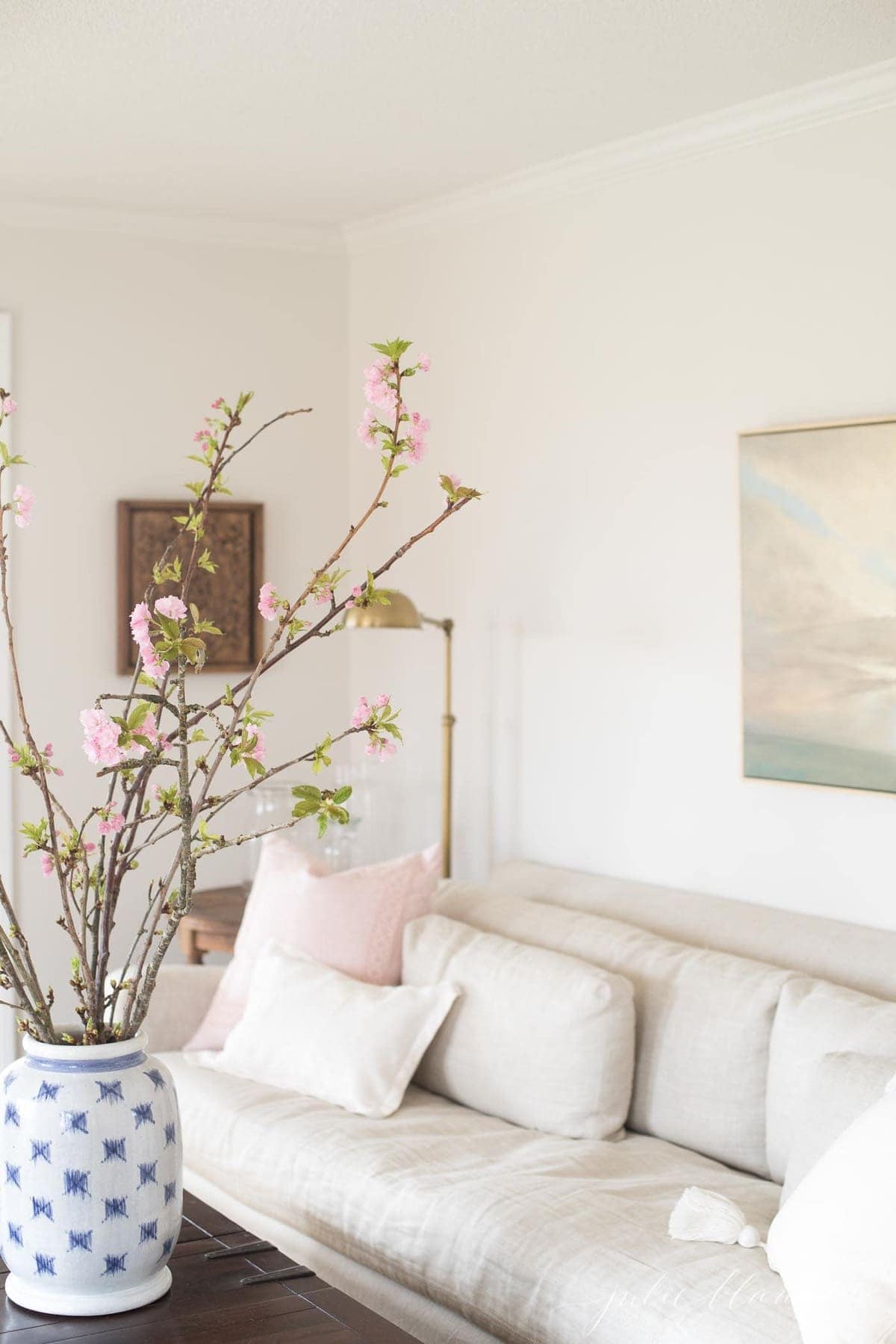 A neutral living room with a linen sofa and tall pink branches in a blue and white vase.