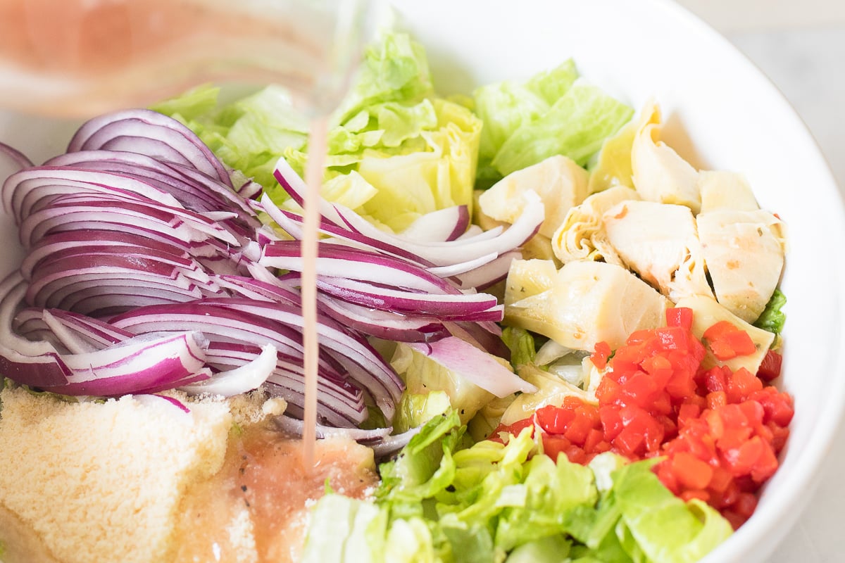 Fresh ingredients for an Italian salad including lettuce, red onions, chicken, tomatoes, and grated cheese in a bowl.