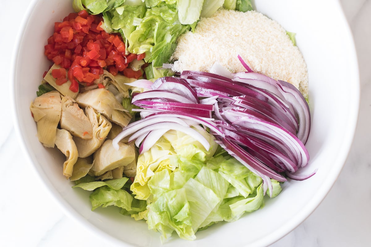 Bowl of Italian salad ingredients including lettuce, tomatoes, artichokes, red onions, and a slice of bread.