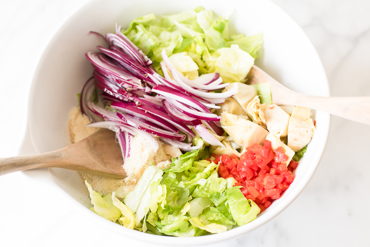 A salad bowl with chopped lettuce, sliced red onions, diced tomatoes, and chunks of chicken, accompanied by two wooden serving spoons and Italian salad dressing.