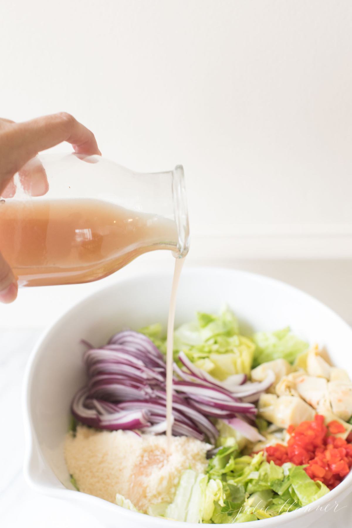 A bottle of red wine vinaigrette pouring over an italian salad. 