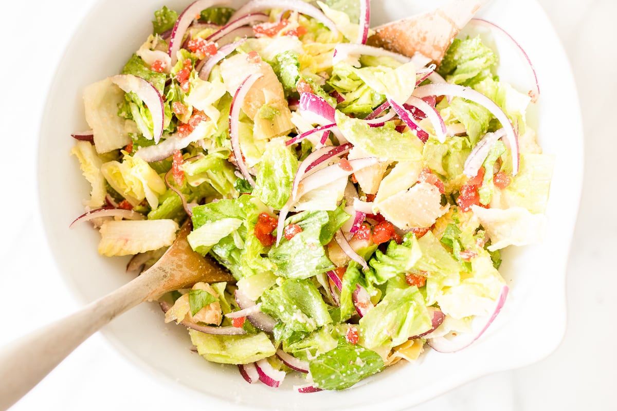 A fresh Italian salad mixed with lettuce, red onions, and tomatoes, partially tossed with wooden utensils.