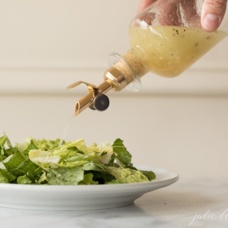 White wine vinaigrette in a glass bottle with a gold spout, poured over a green salad on a white plate.