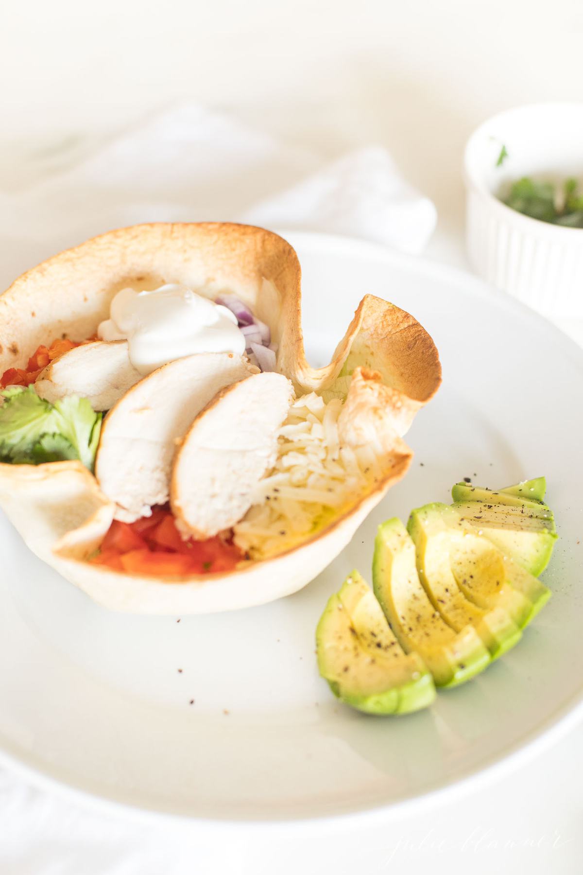 A tortilla bowl full of chicken, cheese, lettuce and more, on a white plate