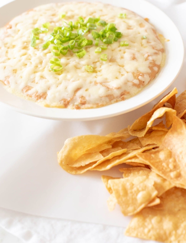 A white bowl full of cream cheese bean dip, topped with diced chives and tortilla chips to the side