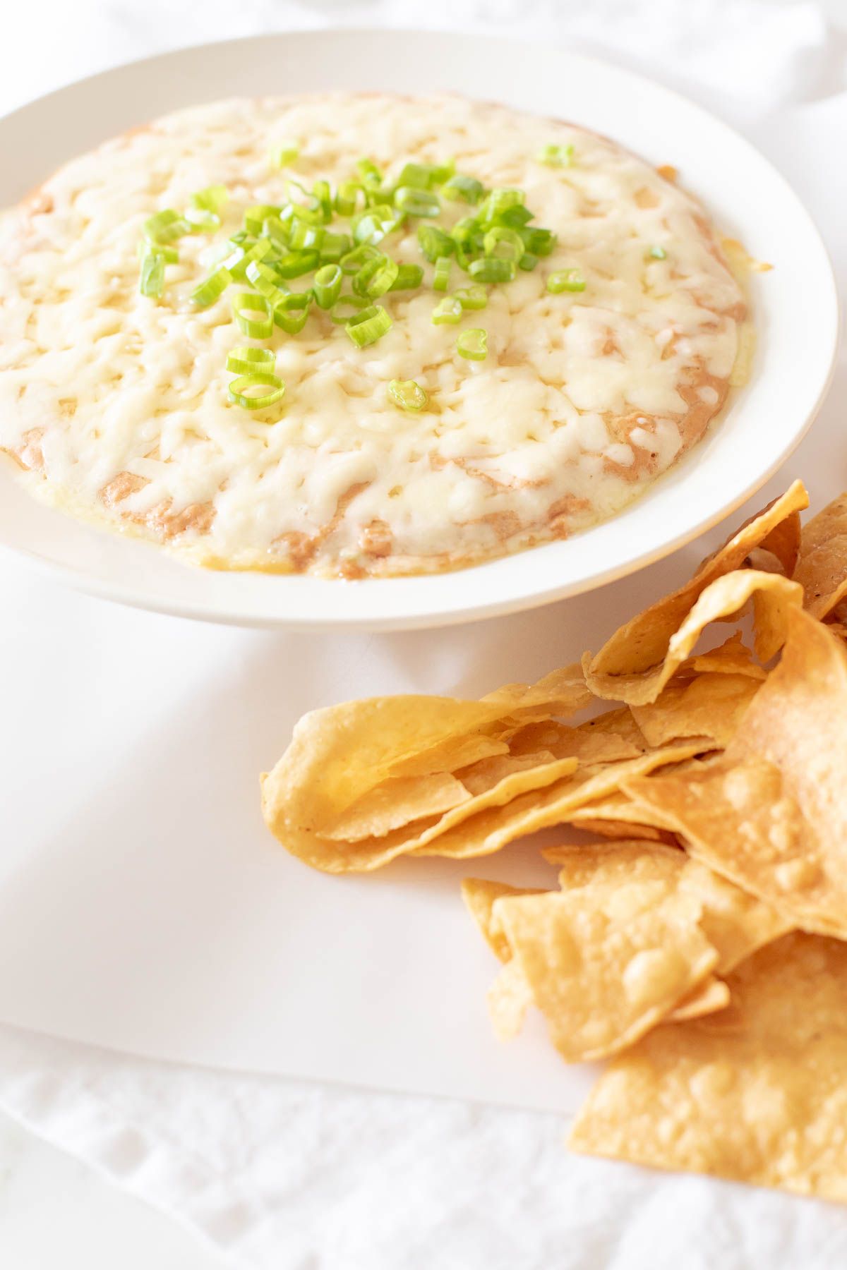 A white bowl full of cream cheese bean dip, topped with diced chives and tortilla chips to the side