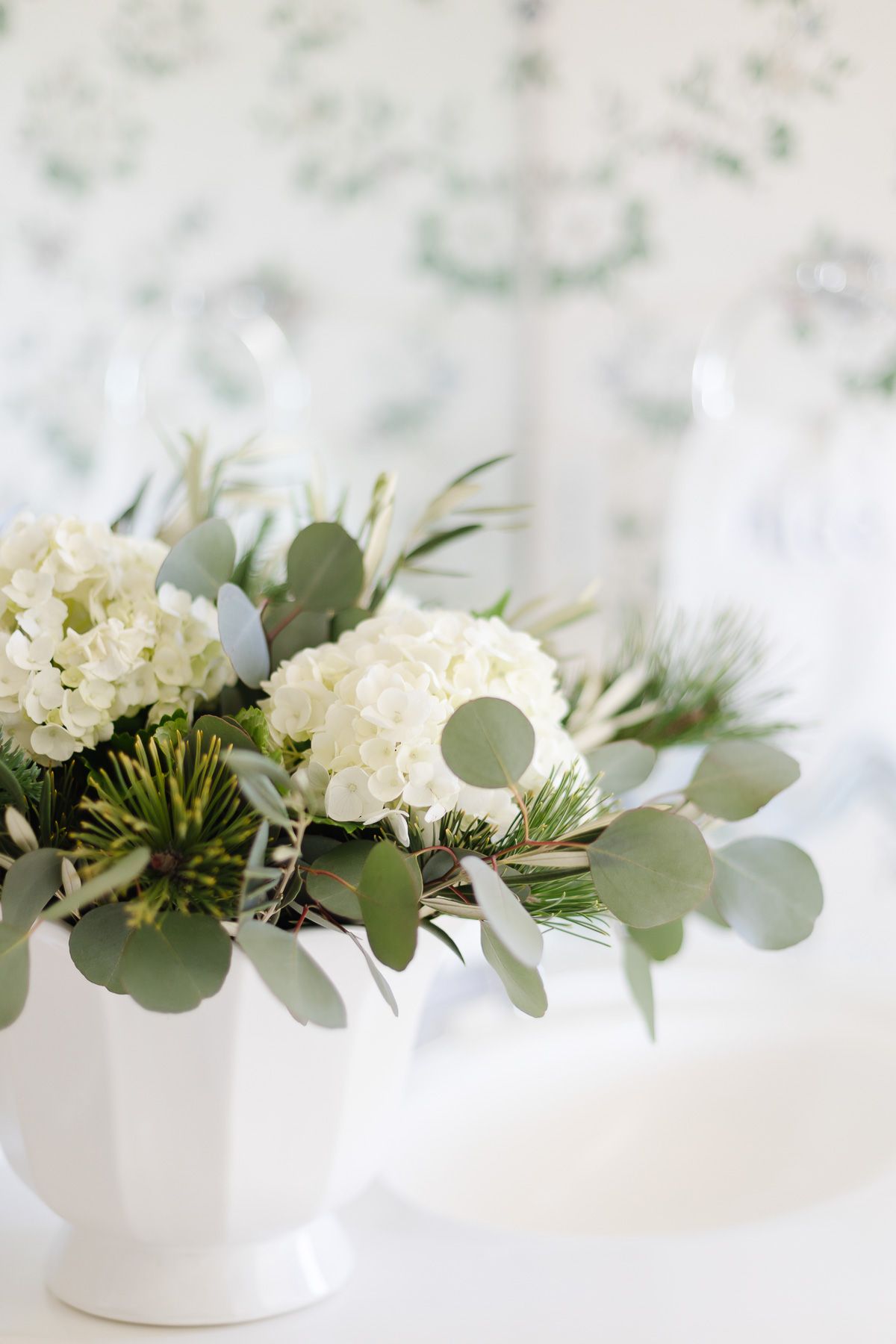 White hydrangeas in a ruffled white ceramic flower vase in a wallpapered bathroom.