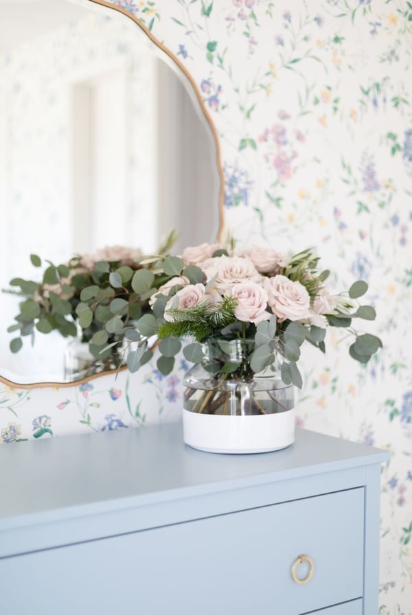 Pink roses in a white dipped flower vase in a wallpapered bedroom.