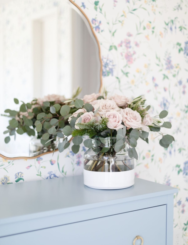 Pink roses in a white dipped flower vase in a wallpapered bedroom.