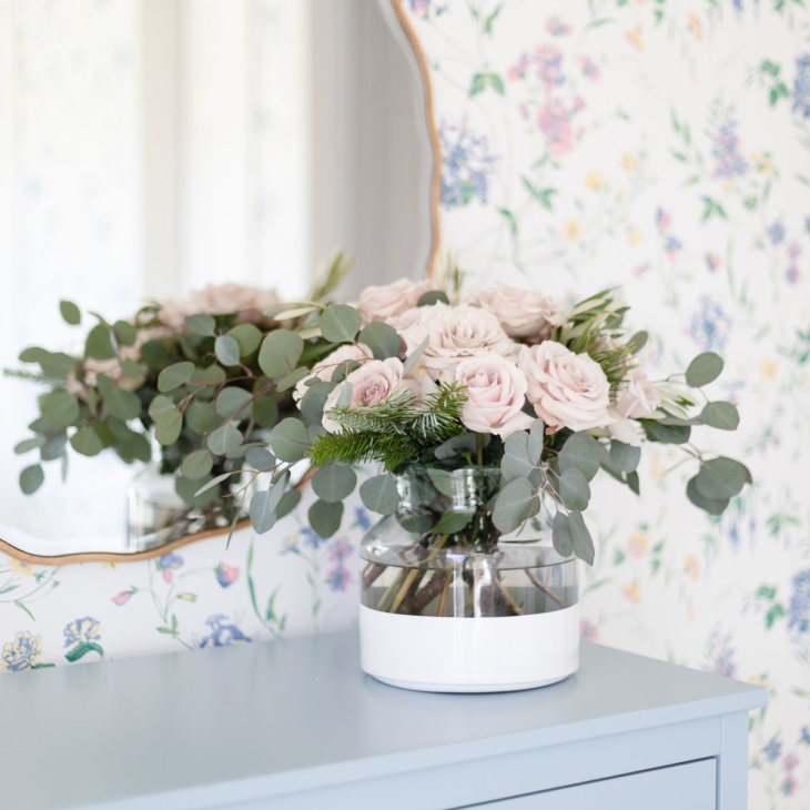 Pink roses in a white dipped flower vase in a wallpapered bedroom.