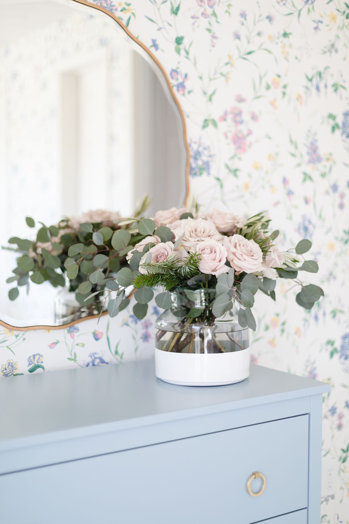 Pink roses in a white dipped flower vase in a wallpapered bedroom. 