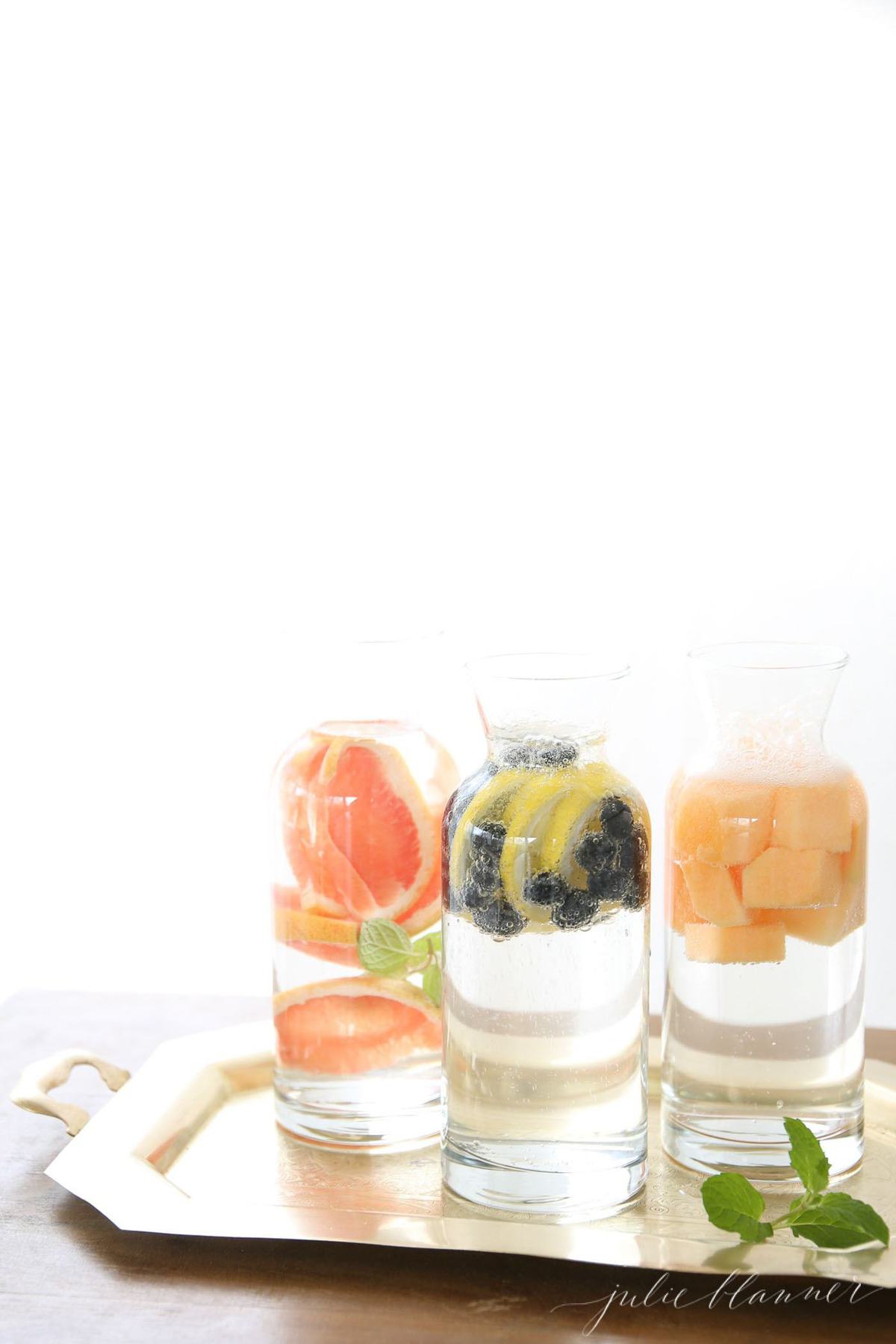 Three glass carafes on a tray, filled with various fruit infused water recipes.
