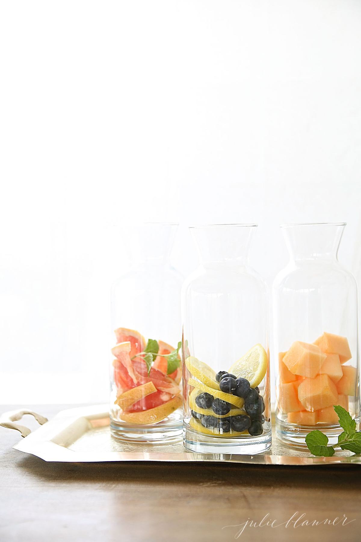 Three glass carafes full of fresh sliced fruit and berries for fruit infused water recipes.