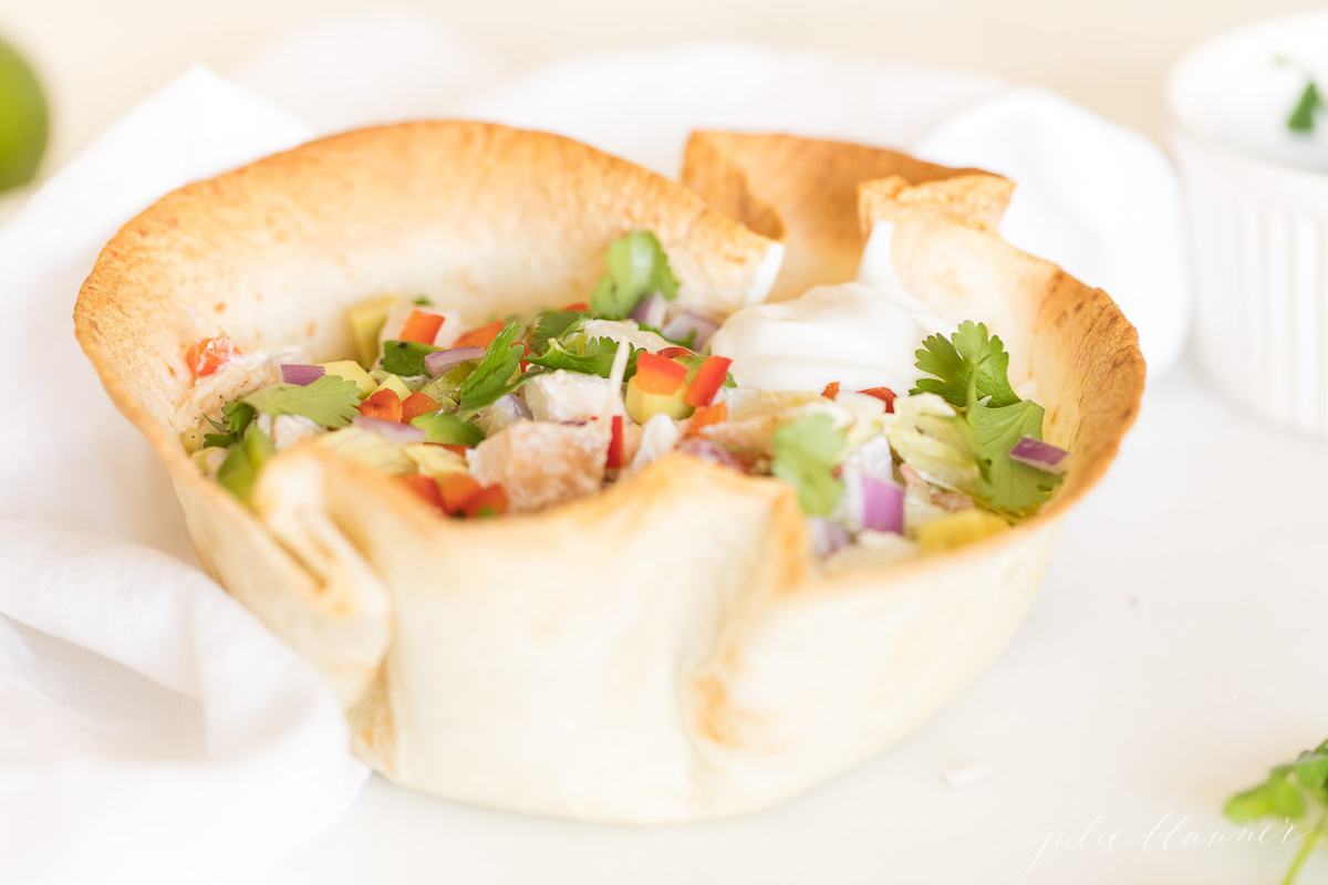 A tortilla bowl filled with chicken, lettuce, vegetables and more, on a white surface.