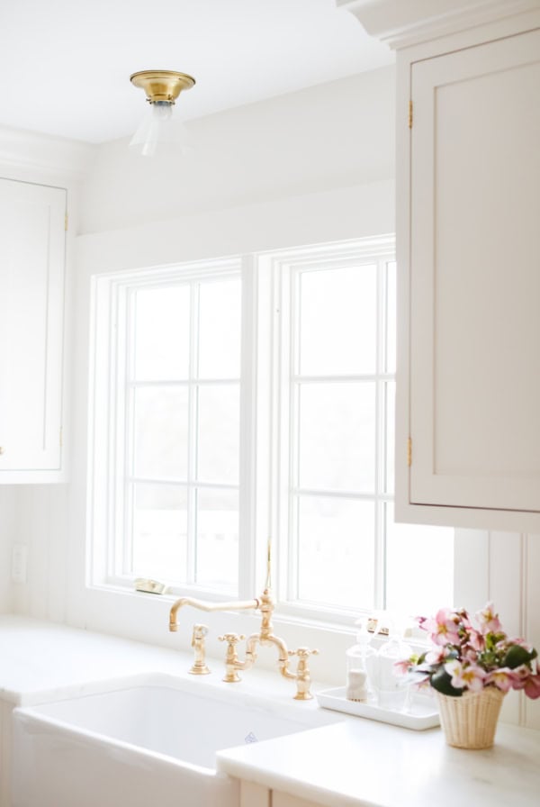 Bright kitchen with white cabinets, a farmhouse sink, brass faucet, and a basket of pink flowers on the counter in front of large windows—perfect for keeping spotless with your favorite marble cleaner.