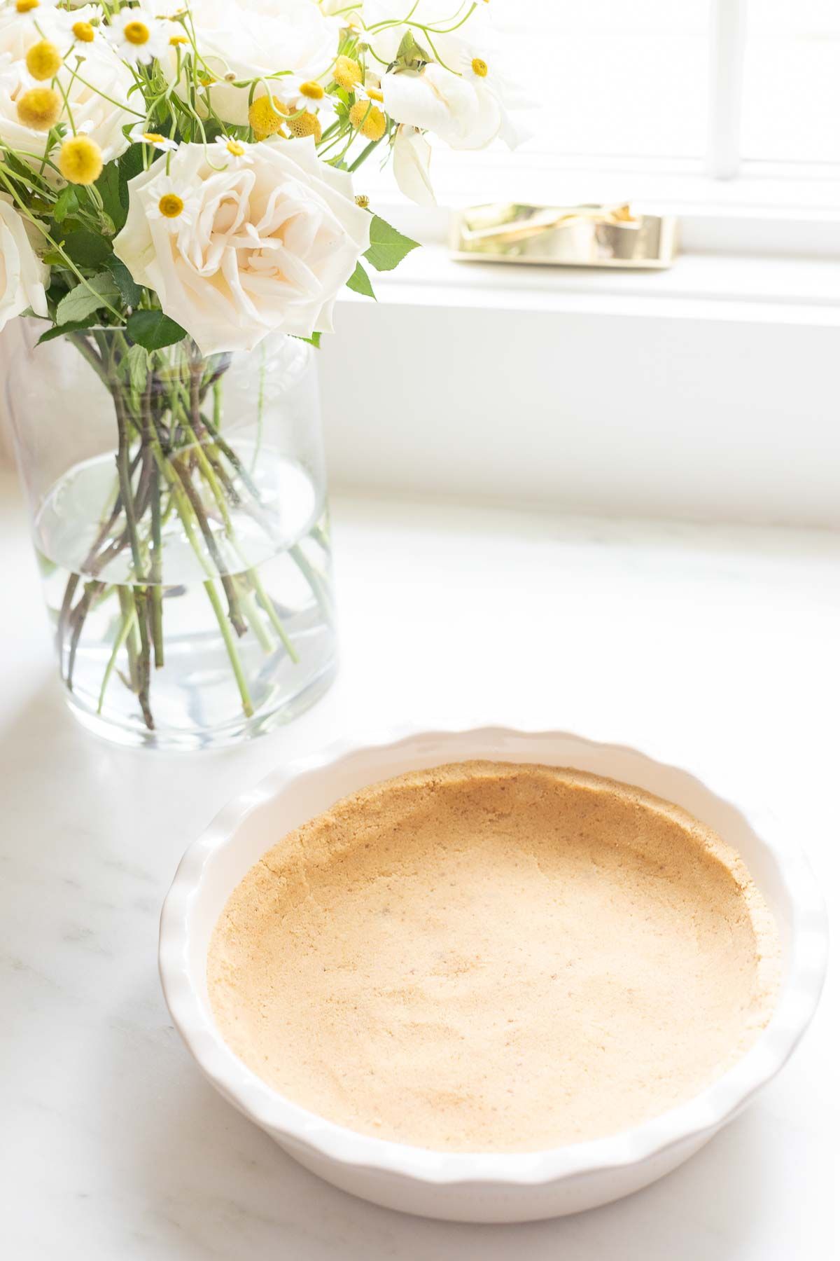 A shortbread crust pressed into a white ruffled ceramic pie dish on a marble surface, flowers to the side
