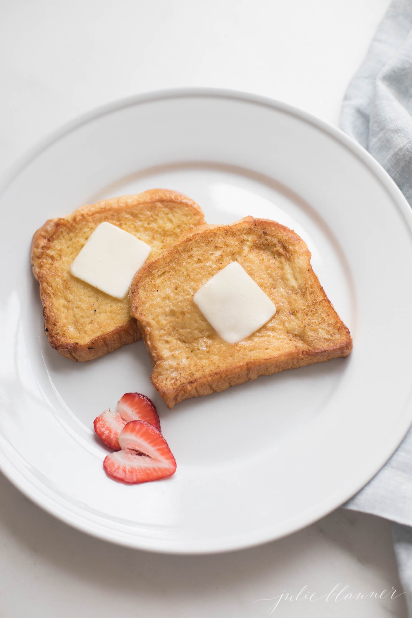 Brioche French toast topped with butter on a white plate, sliced strawberries as garnish.