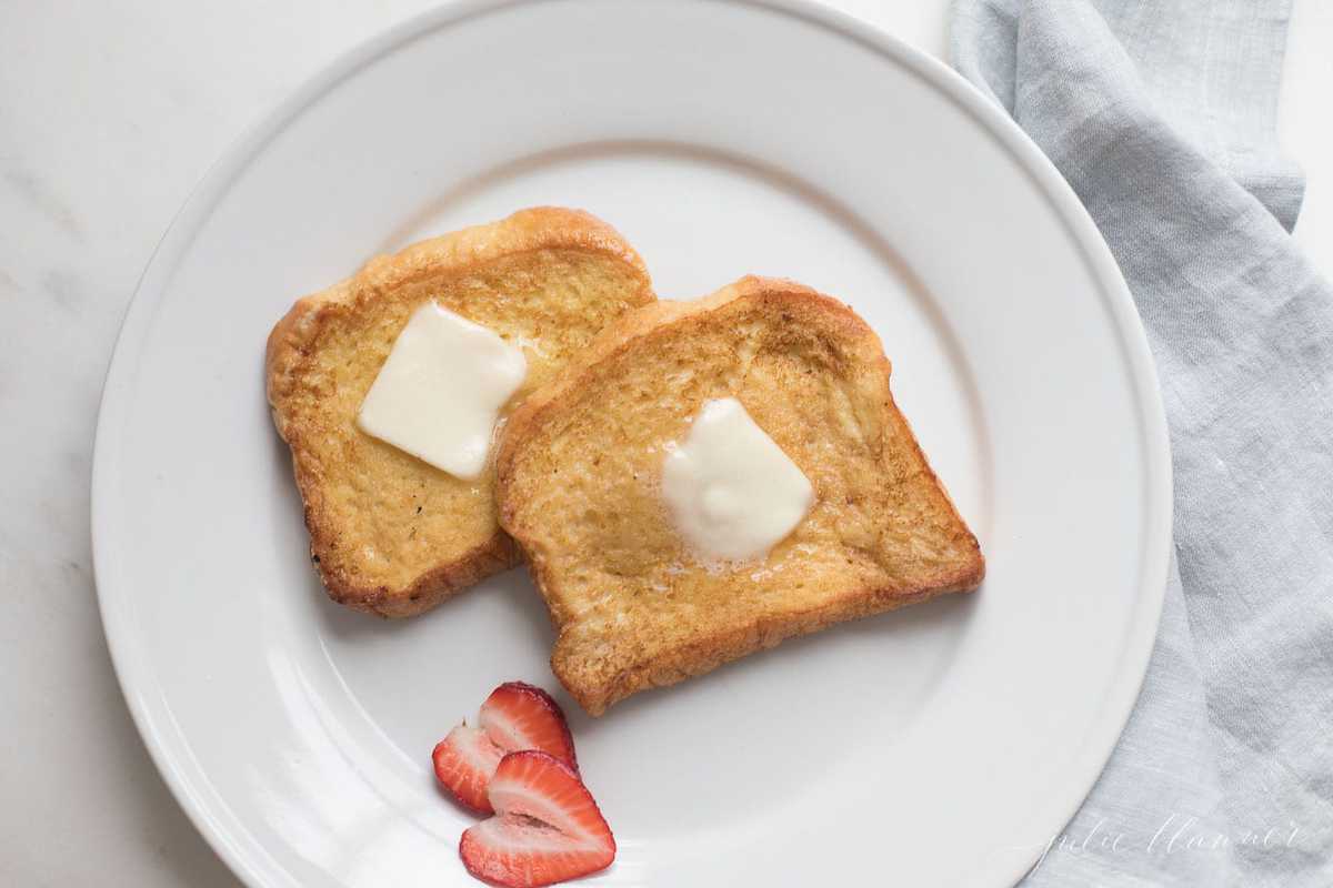 Brioche French toast topped with butter on a white plate, sliced strawberries as garnish.