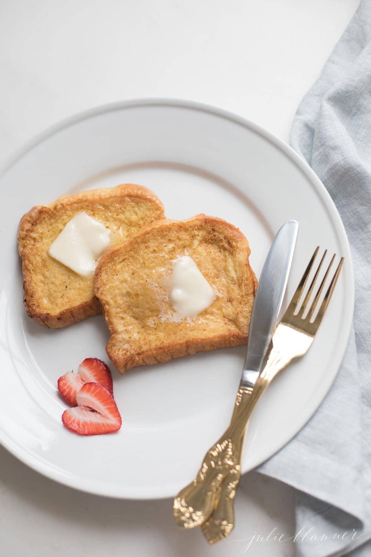 Brioche French toast topped with butter on a white plate, knife and fork to the side.