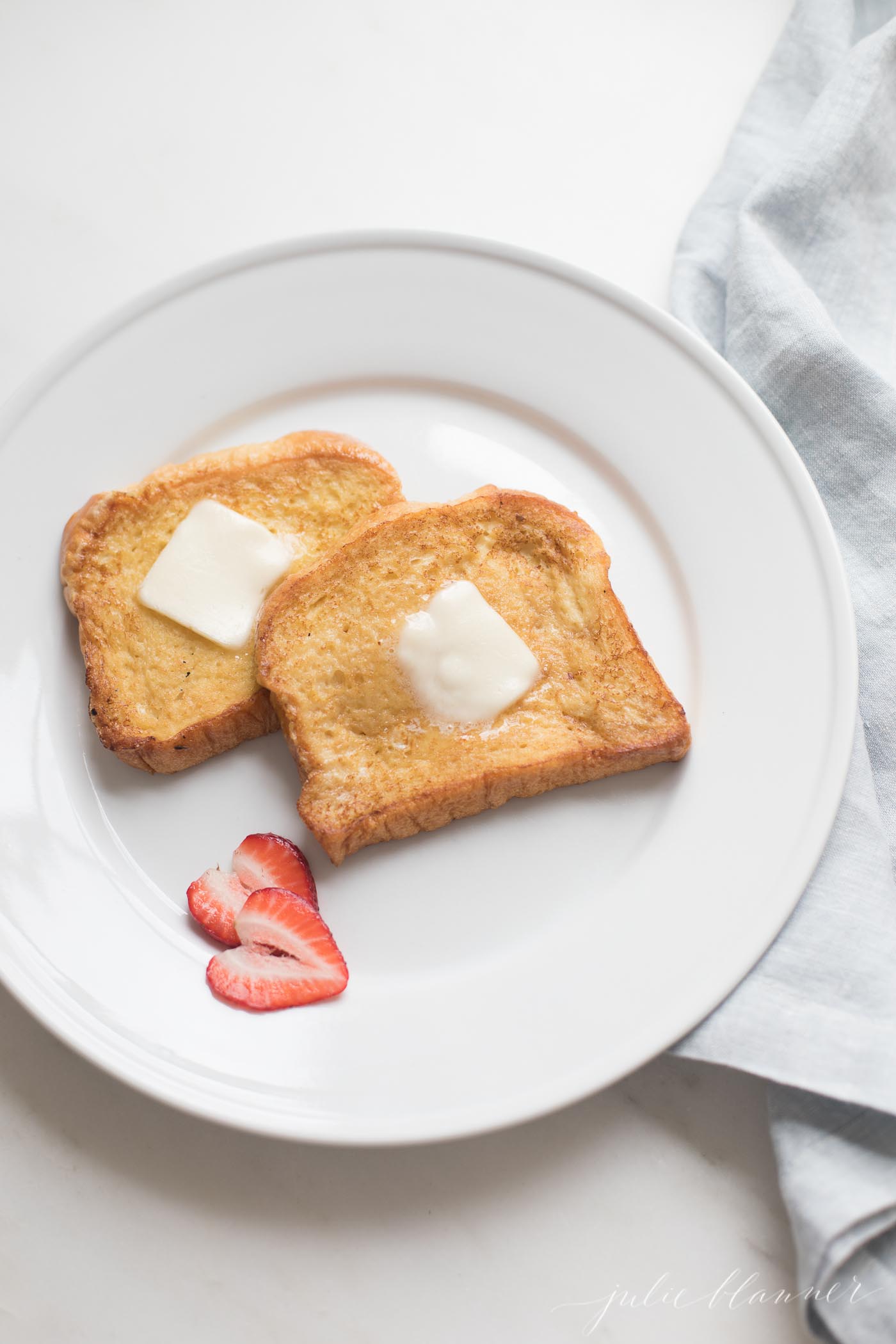 Brioche French toast topped with butter on a white plate, sliced strawberries as garnish.
