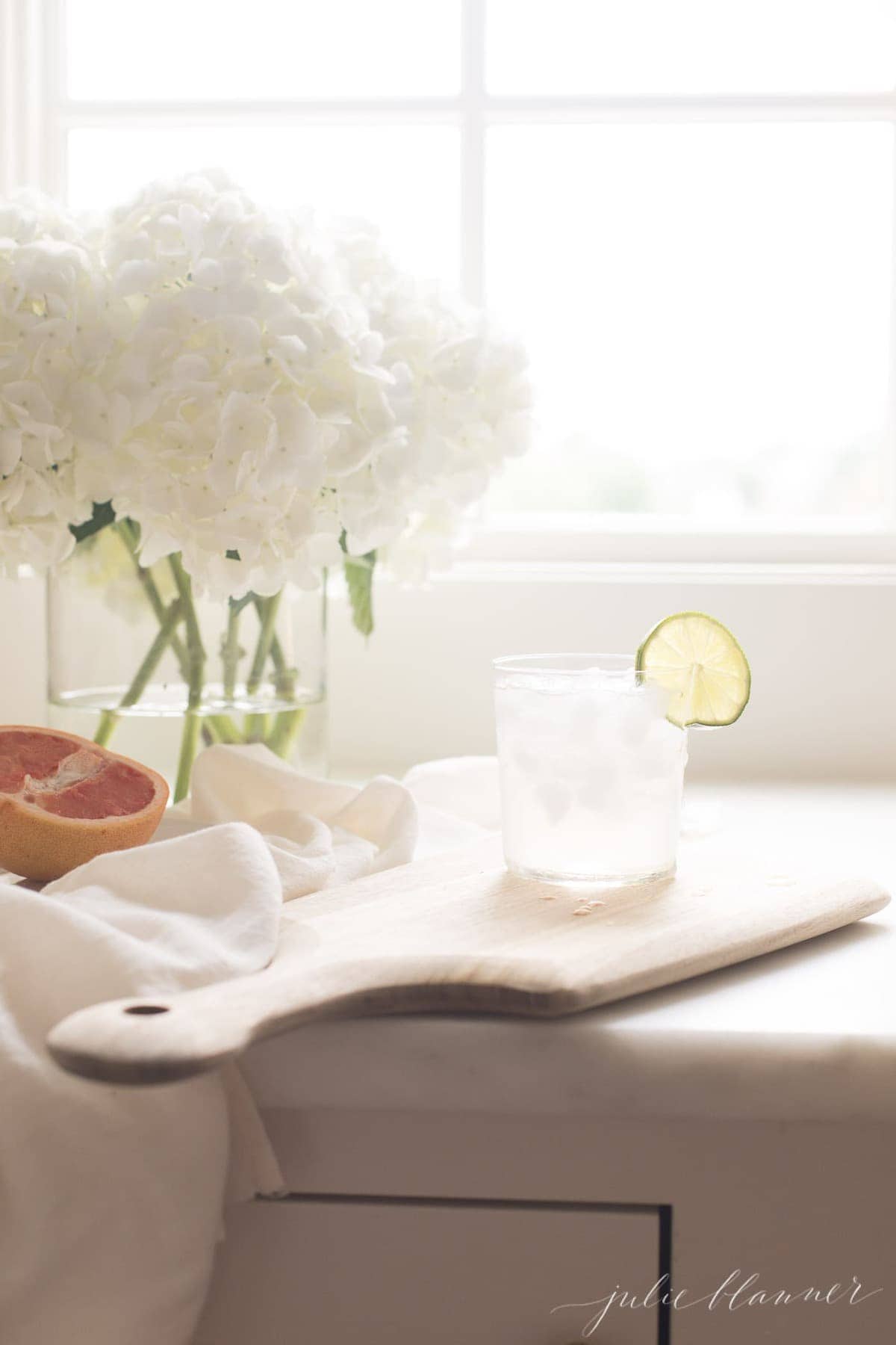 A cutting board with a fresh paloma drink on top, floral arrangement in background.