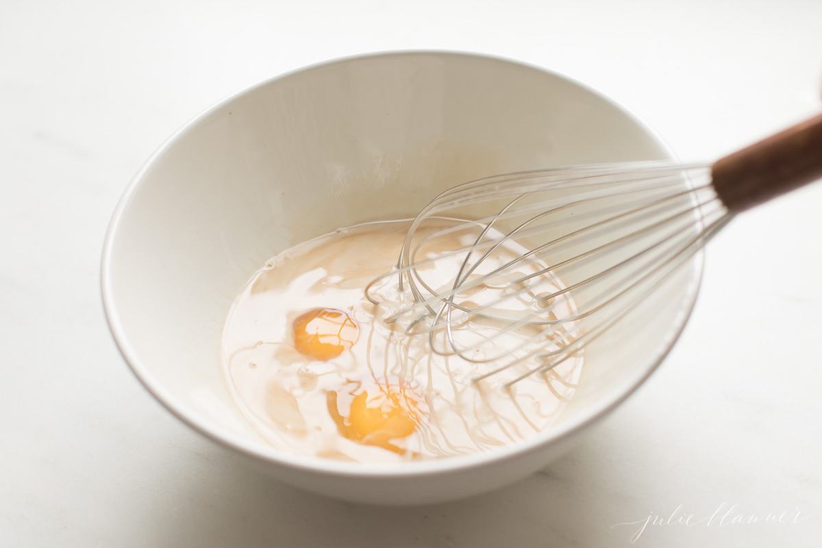 A whisk, whisking eggs and milk in a white bowl on a marble countertop