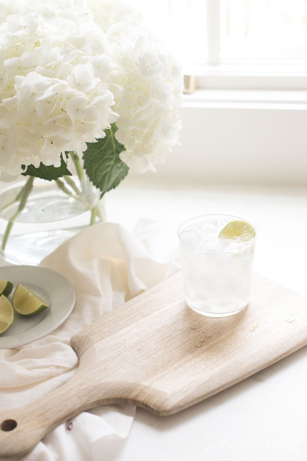 A fresh paloma recipe in a clear glass on ice, with a vase of fresh flowers in the background.