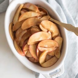 A white oval baking dish filled with baked apple slices, gold spoon coming out the side.