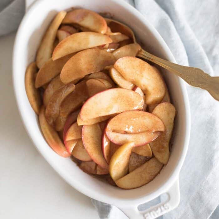 A white oval baking dish filled with baked apple slices, gold spoon coming out the side.