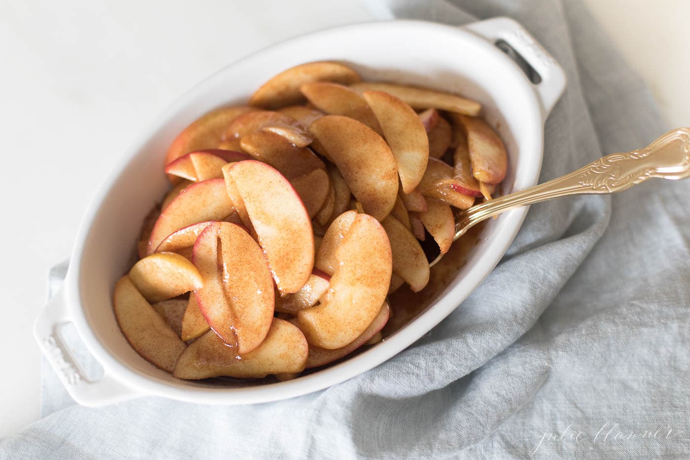 An oval baking dish filled with baked apple slices with a blue towel in the background. 