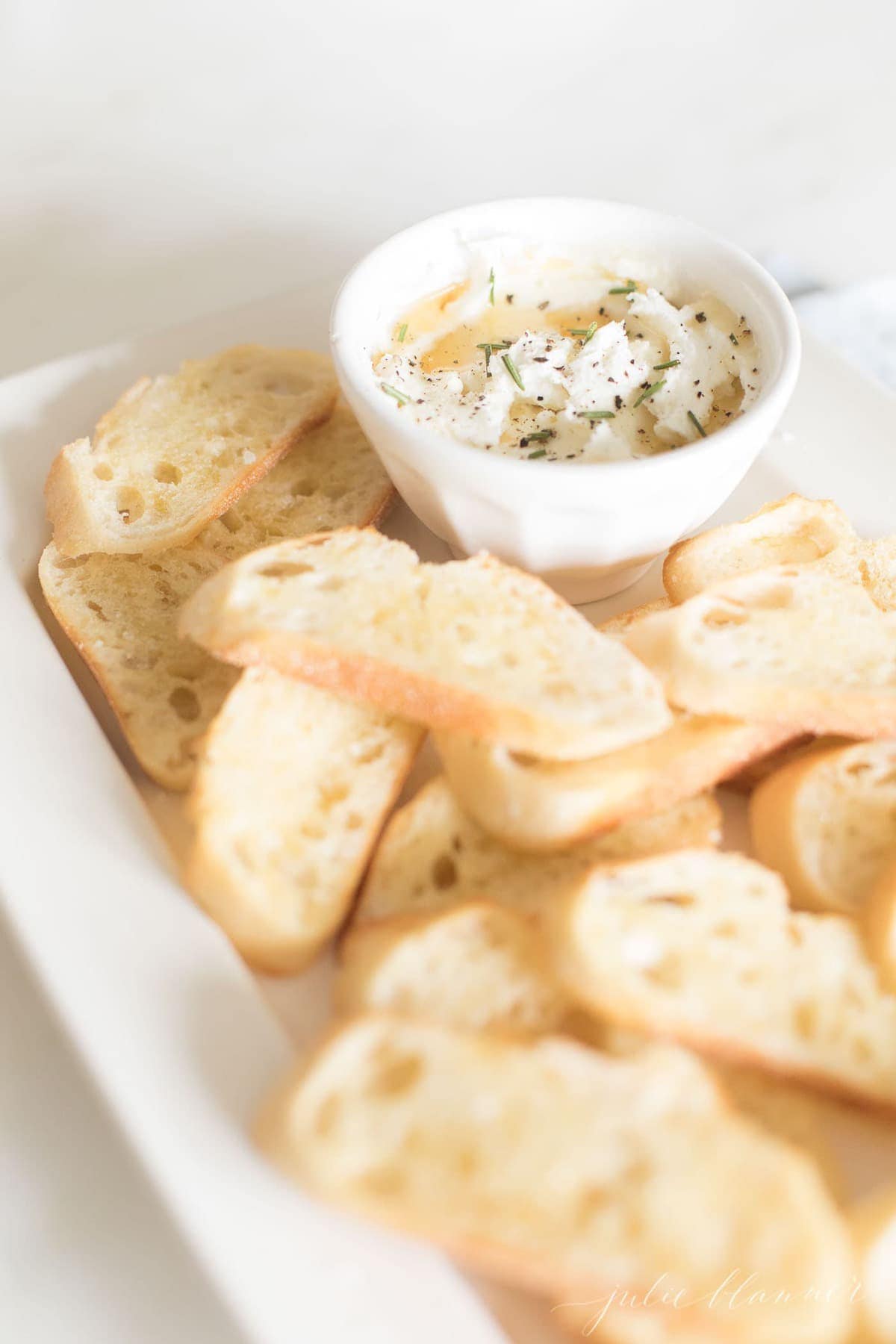 Whipped goat cheese spread in a white bowl on a white platter full of sliced crostini.