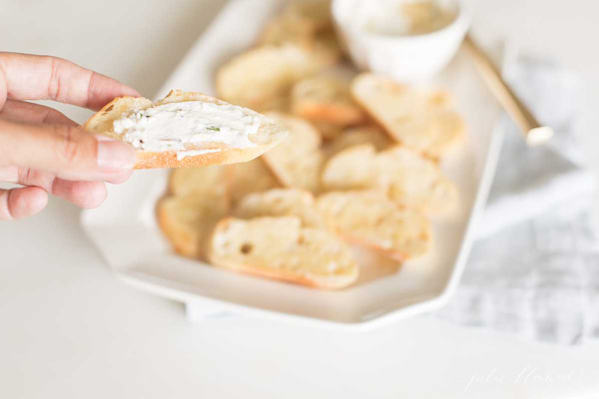 A hand holding a slice of crostini topped with whipped goat cheese, platter of crostini in the background.